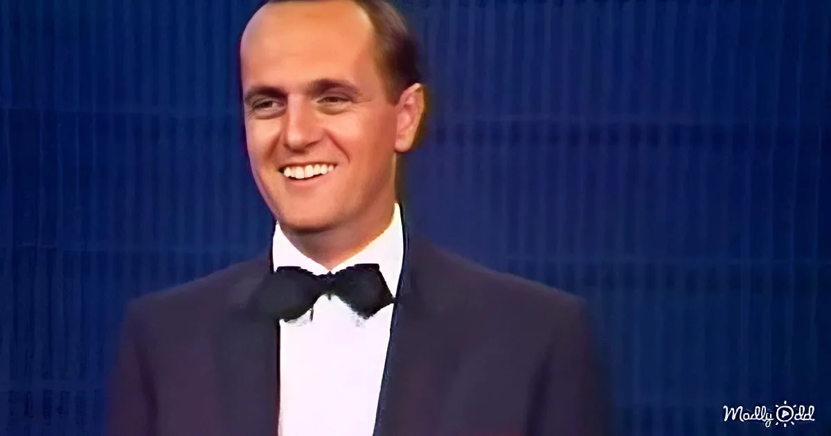 A man in a tuxedo smiles brightly against a blue background.