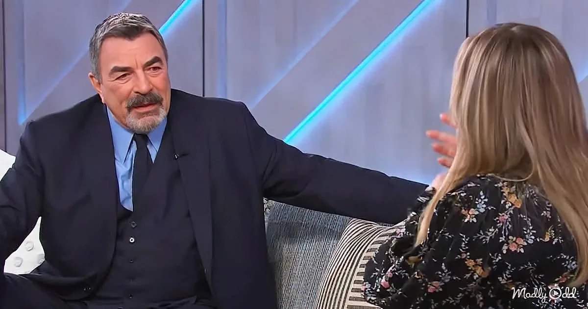 A man in a suit extends his arm towards a woman with long blonde hair, both seated on a couch.