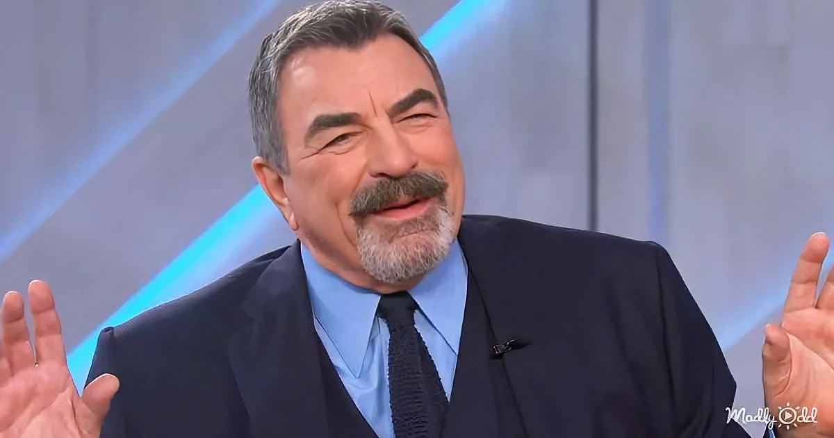 A man in a suit and tie gestures with both hands, appearing to speak or explain something.