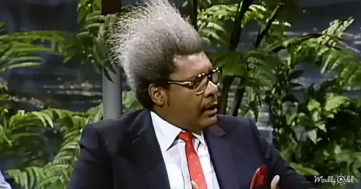 A man in a suit and red tie, with a distinctive hairstyle, is speaking in front of green foliage.