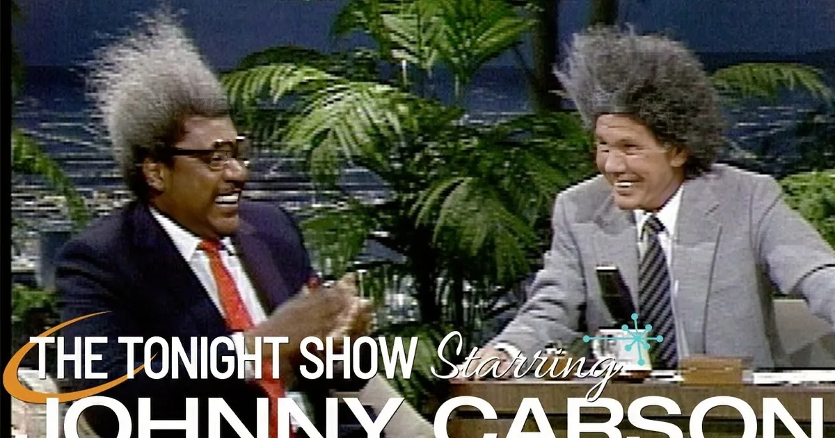 Two men in suits, one with wild hair, are laughing on a talk show set with a palm tree backdrop.