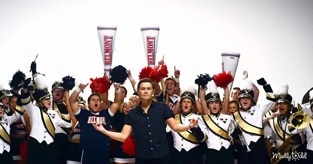A man in a black shirt stands in front of a group of enthusiastic band and cheerleaders, all wearing Belmont uniforms.