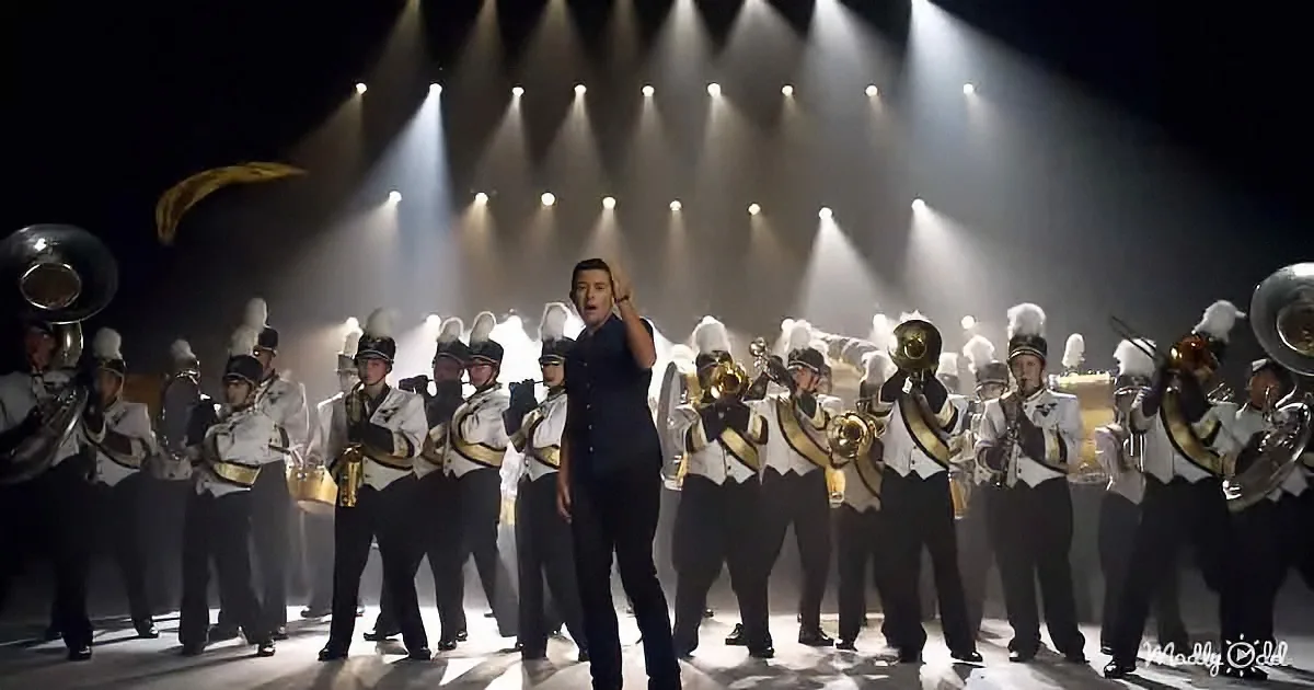 A man in a black suit stands center stage, conducting a marching band under bright stage lights.