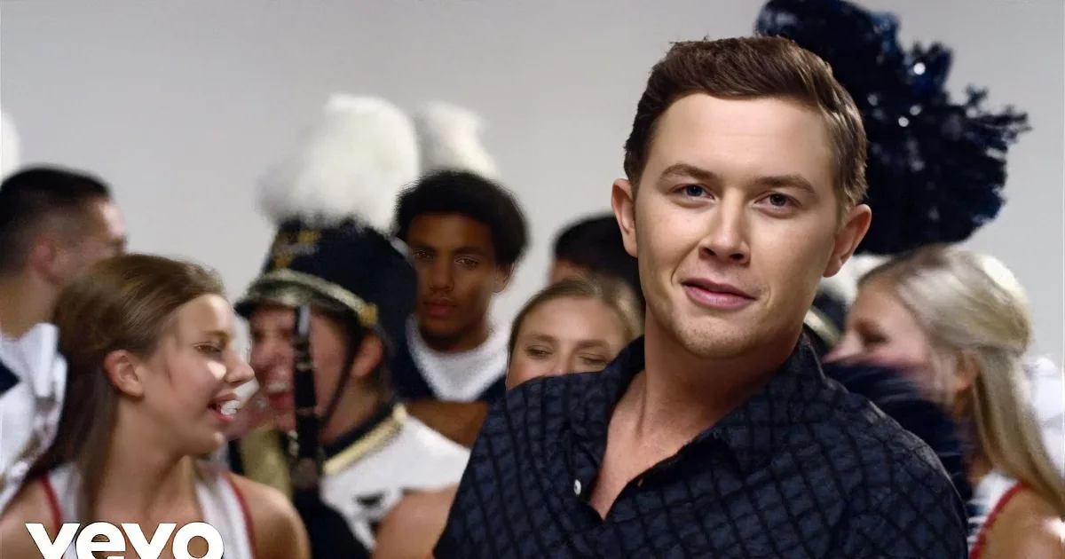 A man in a dark shirt smiles in front of a group of cheerleaders and band members.