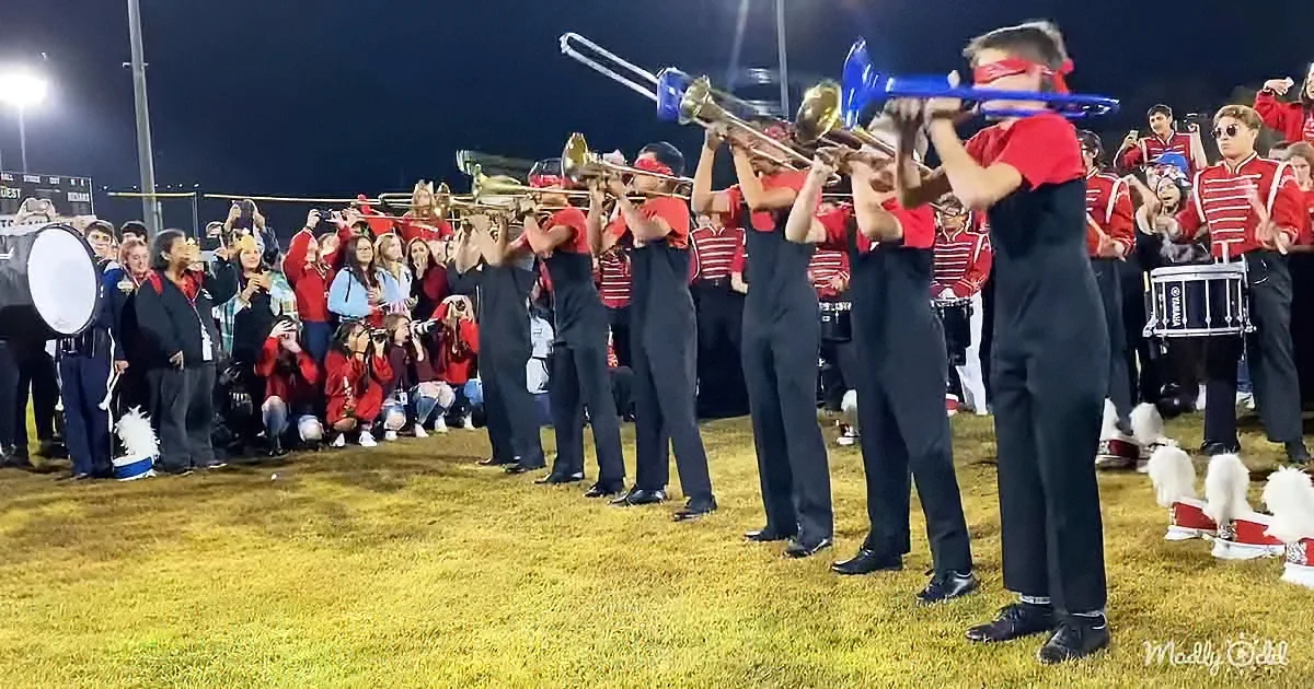 A marching band in red and black uniforms plays brass instruments on a field at night, with spectators cheering.