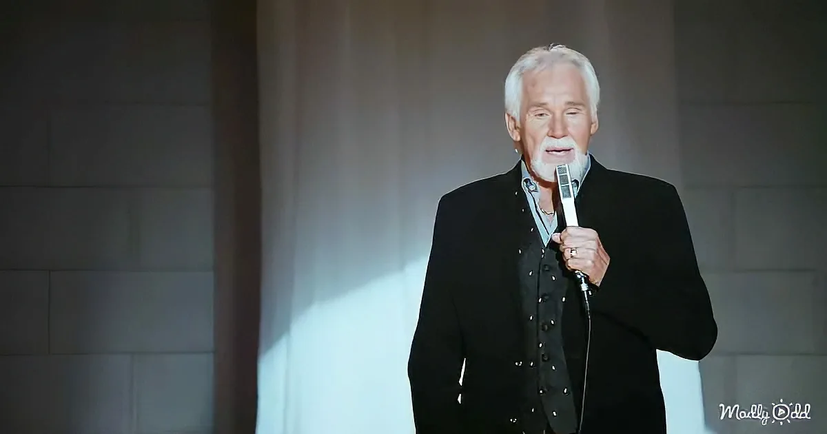 A man in a black suit holds a microphone on stage, illuminated by a spotlight.