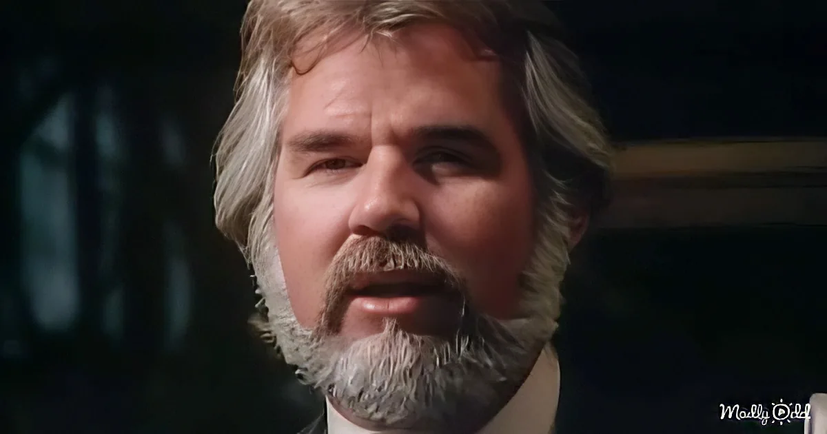 A man with gray hair and a beard, wearing a suit, is looking to the side in a dimly lit setting.
