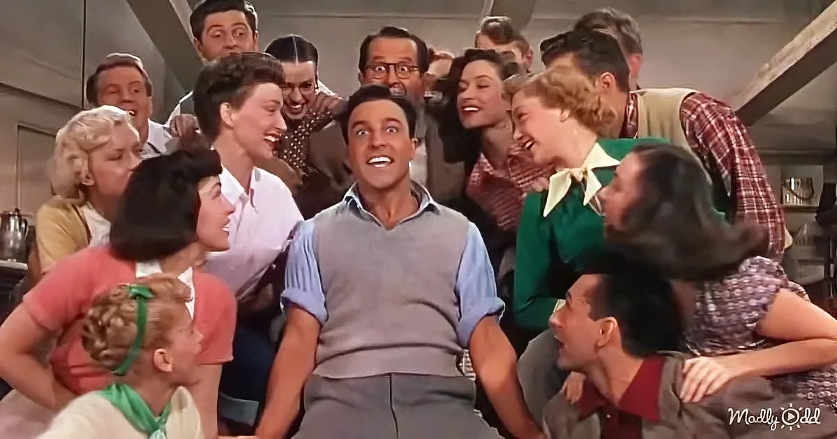 A man in a gray vest and blue shirt is surrounded by a group of people laughing and smiling in a lively scene.