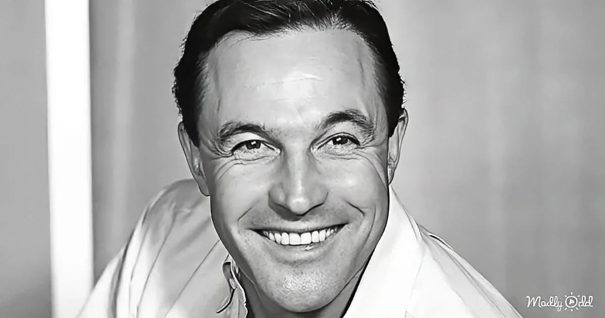 A smiling man with short dark hair, wearing a light-colored shirt, against a blurred background.