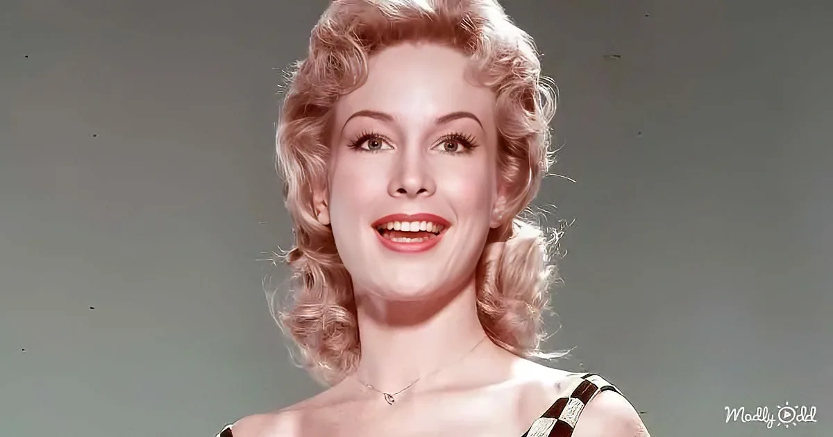 A woman with blonde, curly hair and red lipstick smiles warmly, wearing a necklace and a patterned top.