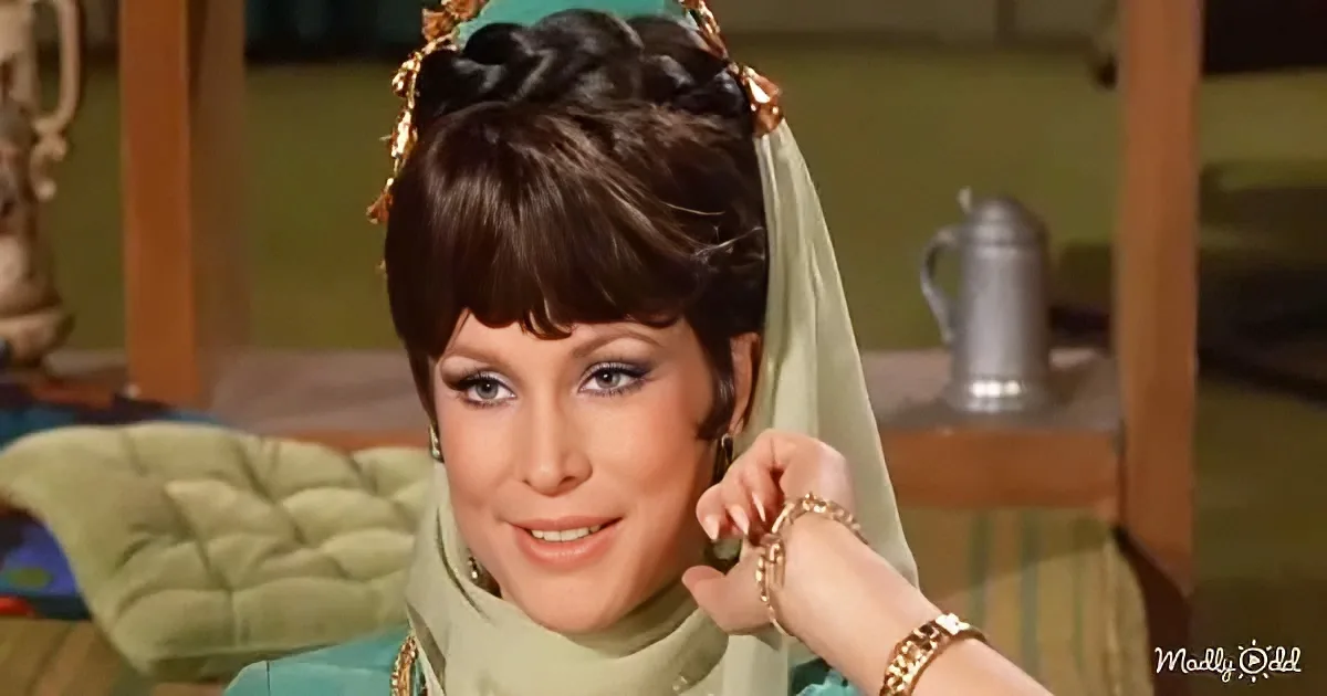 A woman with dark hair styled in a bun, wearing a green outfit and accessories, smiles while holding her hand near her face.
