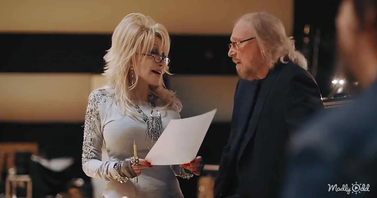 Dolly Parton holds a sheet of paper, engaged in conversation with a man with long hair.