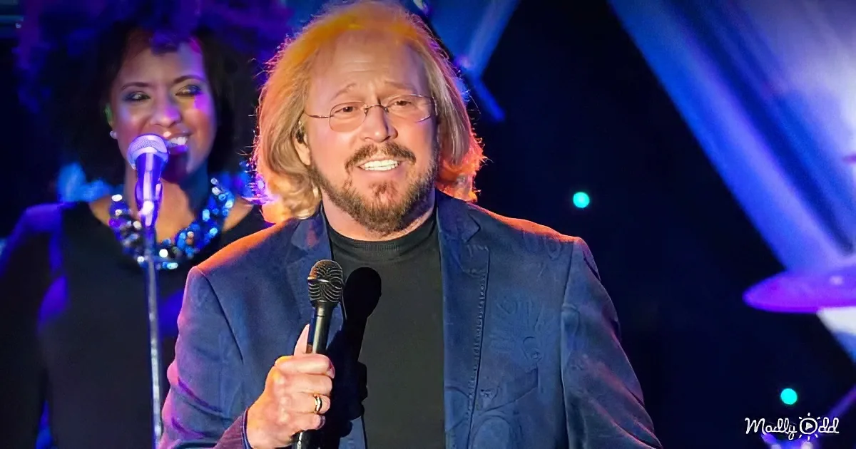 A man with long hair and glasses holds a microphone, singing on stage with a band.