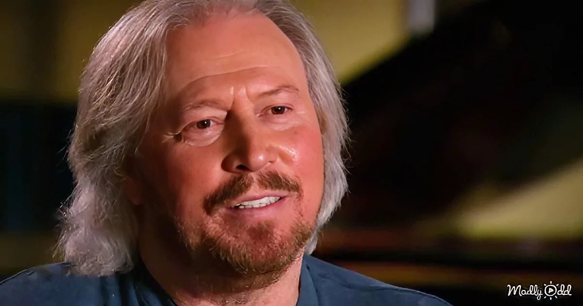 A man with long, light brown hair and a beard, wearing a dark shirt, smiles softly.