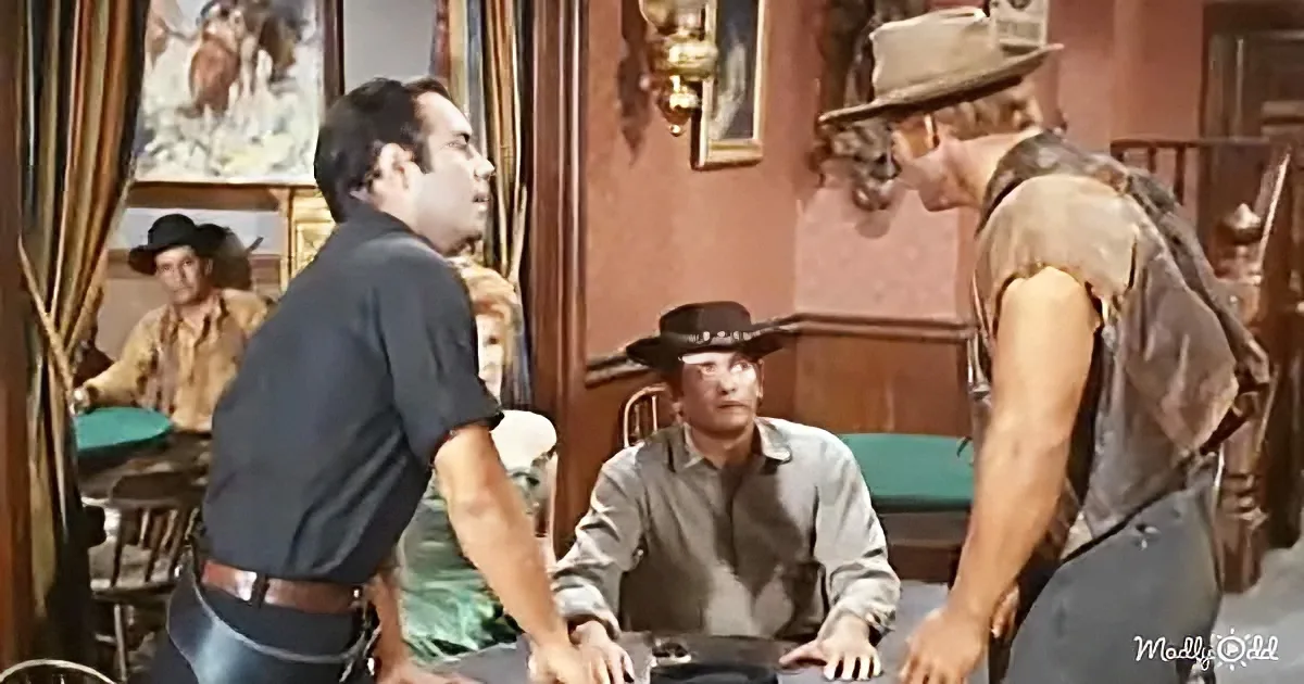 A man in a cowboy hat and vest stands facing another man in a saloon, with a seated man in a hat and shirt in the background.