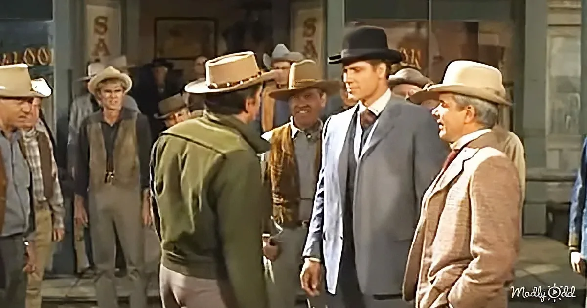 A man in a suit and hat stands among a group of men in cowboy attire in a Western setting.