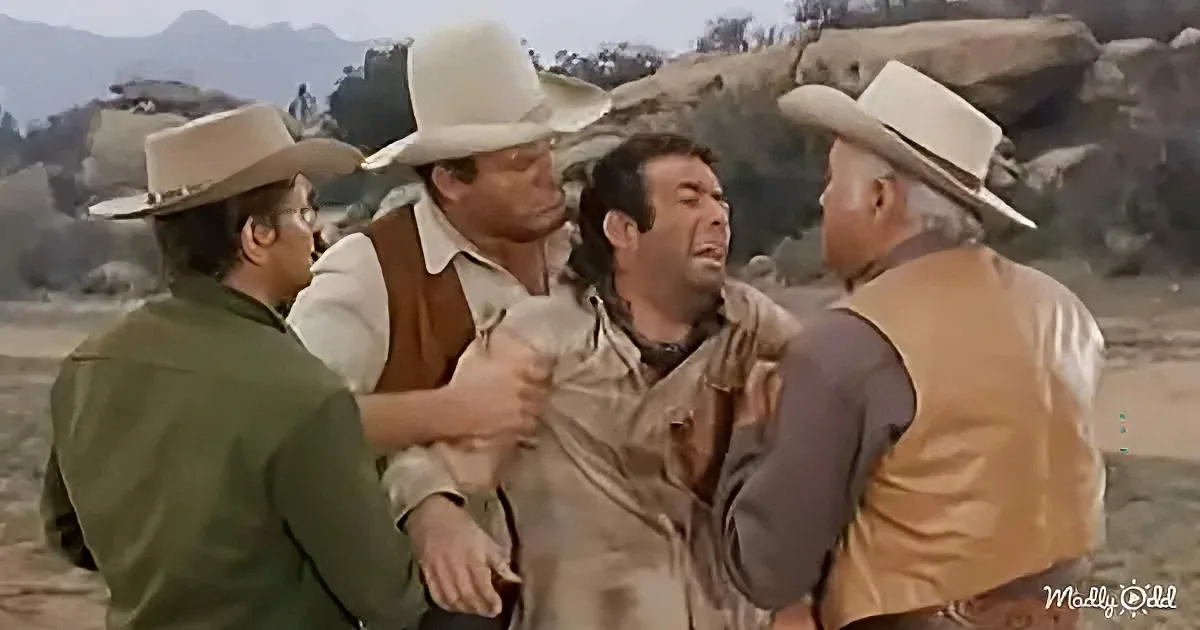 A man in a tan shirt is being restrained by three men in cowboy hats in a desert setting.