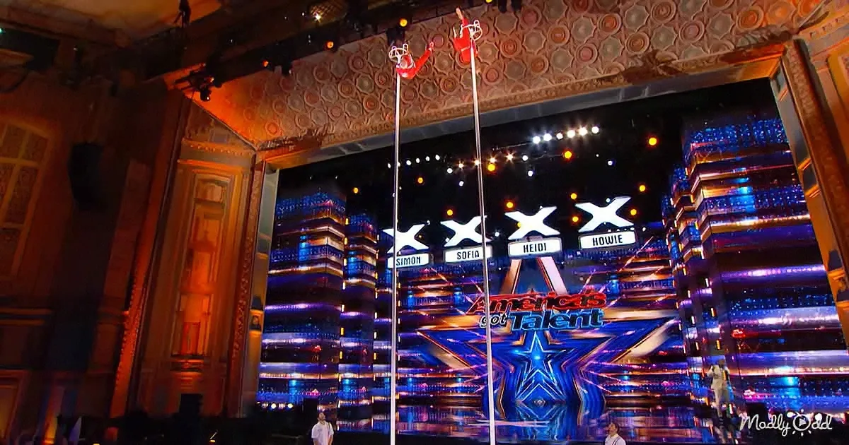 A performer in red attire is suspended on a pole, showcasing an acrobatic routine on a stage with judges' names displayed.