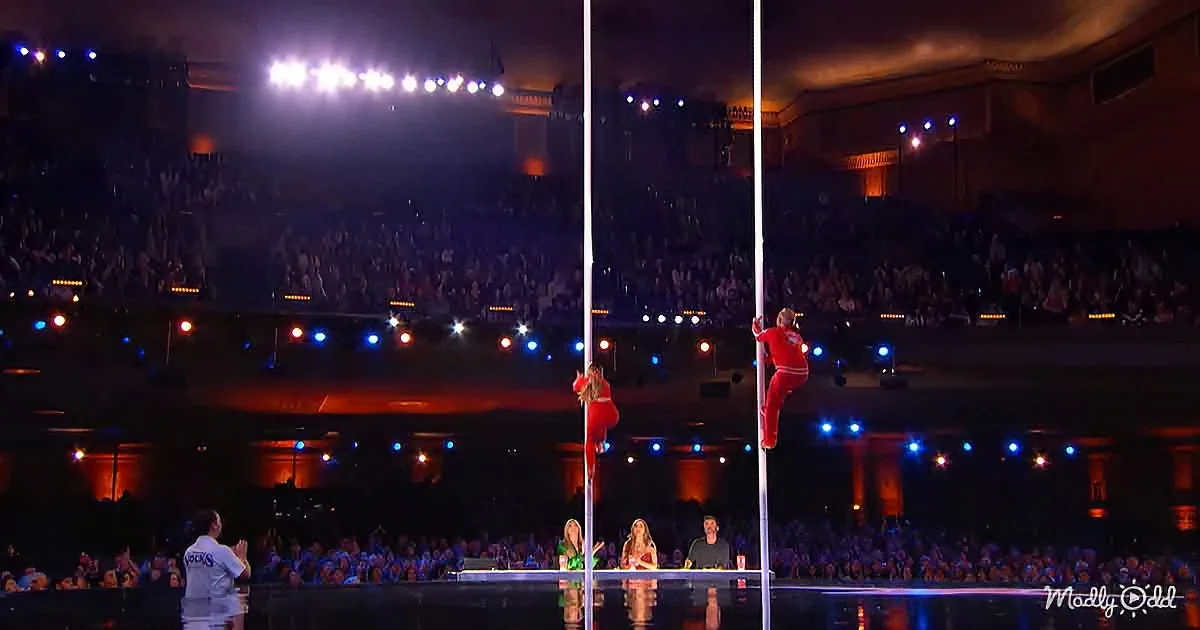 Two pole dancers in red perform on poles in front of a large, cheering audience in a dimly lit arena.