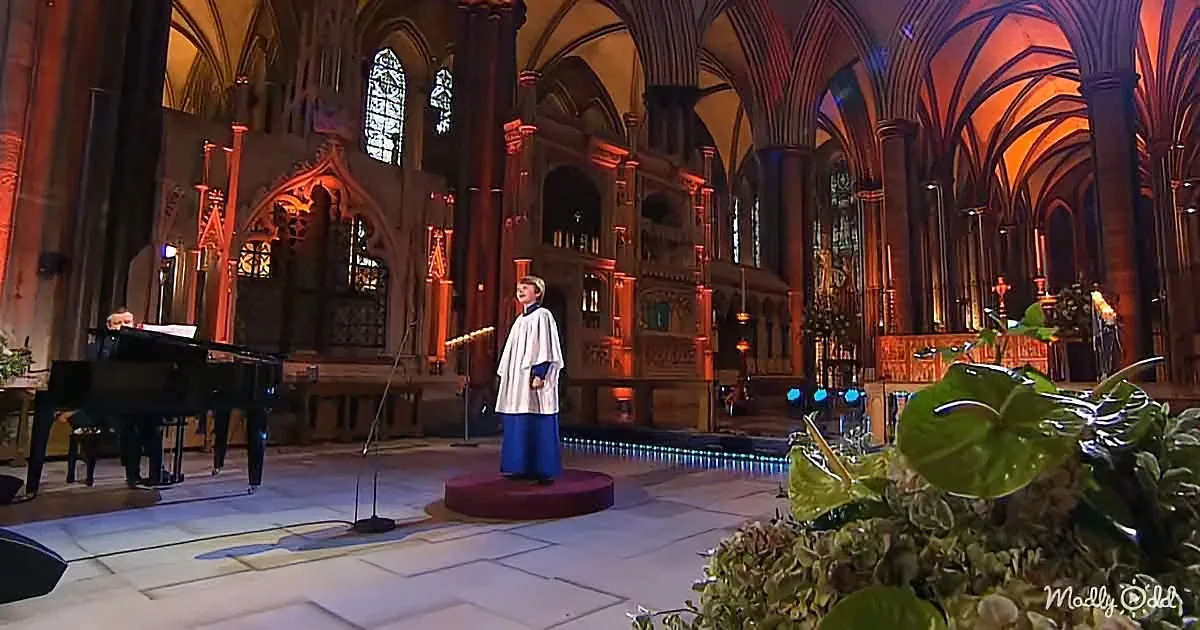 A person in white attire stands on a stage in a grand church, with a pianist and microphone nearby.