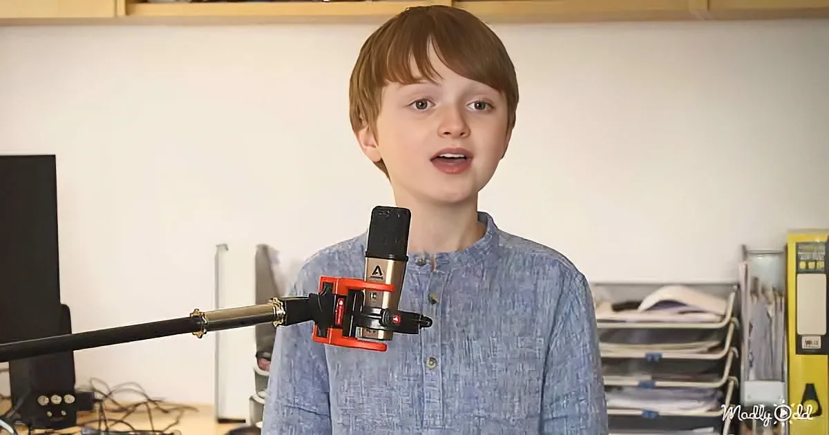 A young boy stands in front of a microphone, speaking in a room with shelves and equipment.