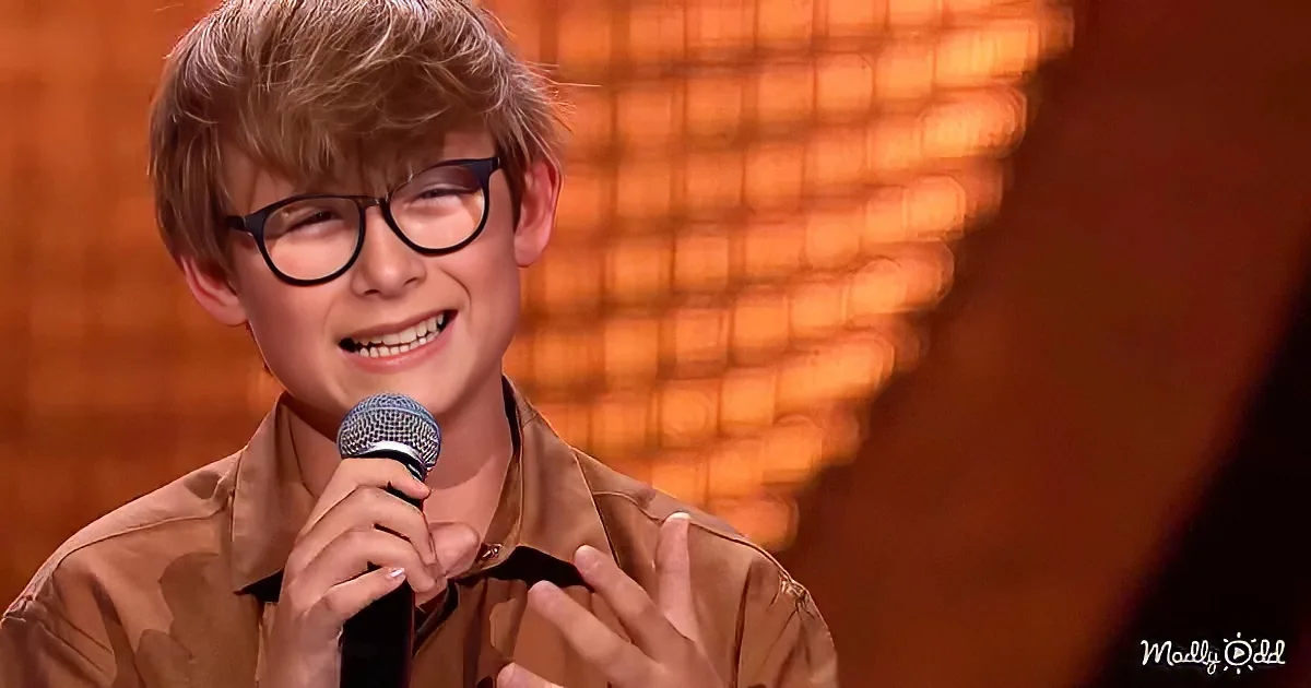 A young boy with glasses smiles while holding a microphone, wearing a brown shirt, against a blurred orange background.