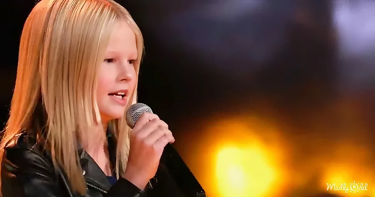 A young person with long blonde hair sings into a microphone on stage with warm lighting.