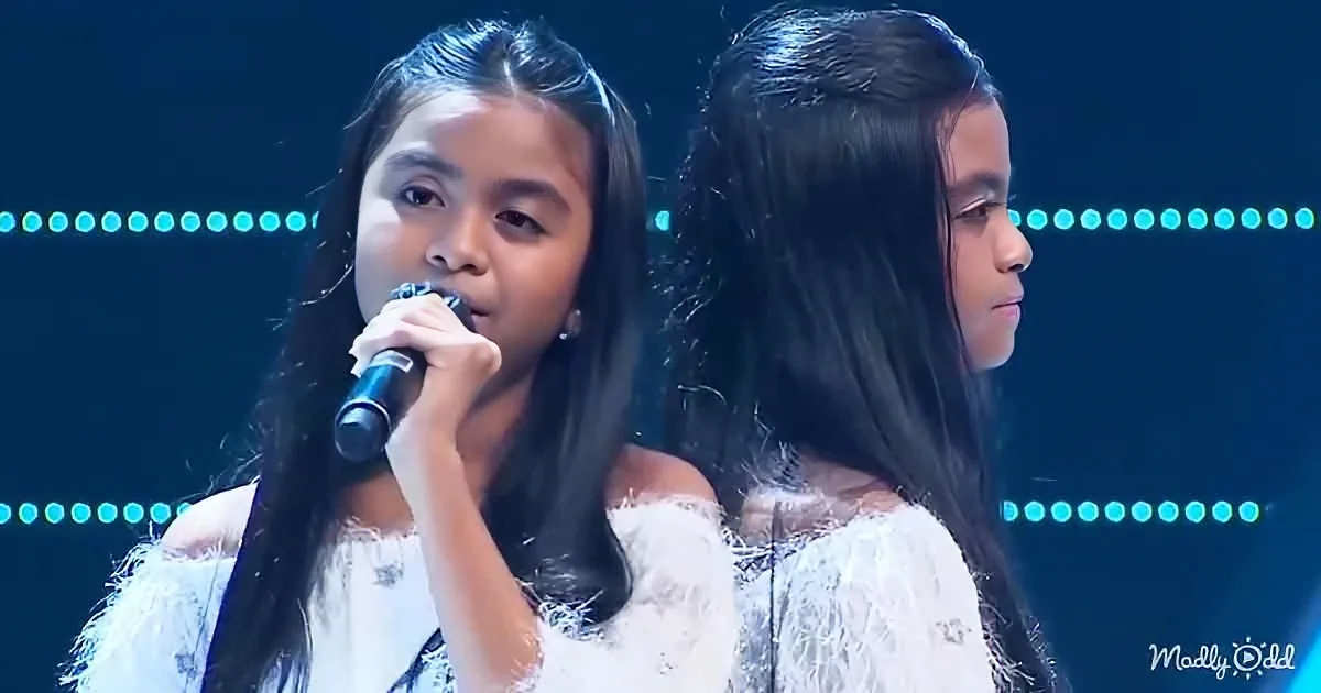 Two young girls in white dresses sing into microphones on a stage with blue lights.