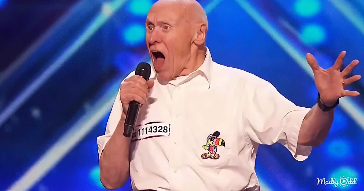 A man in a white shirt with a Mickey Mouse patch and a number on it is singing into a microphone on a stage.