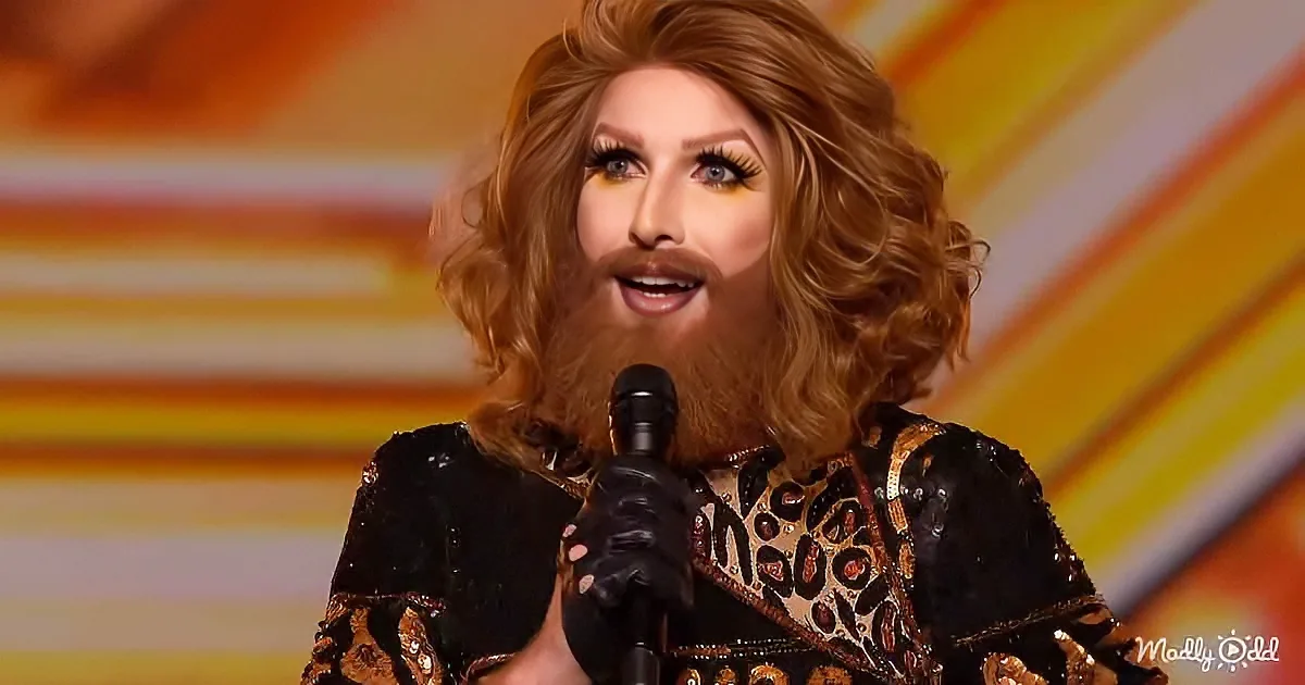 A person with long, curly hair and a beard speaks into a microphone, wearing a leopard-print outfit.