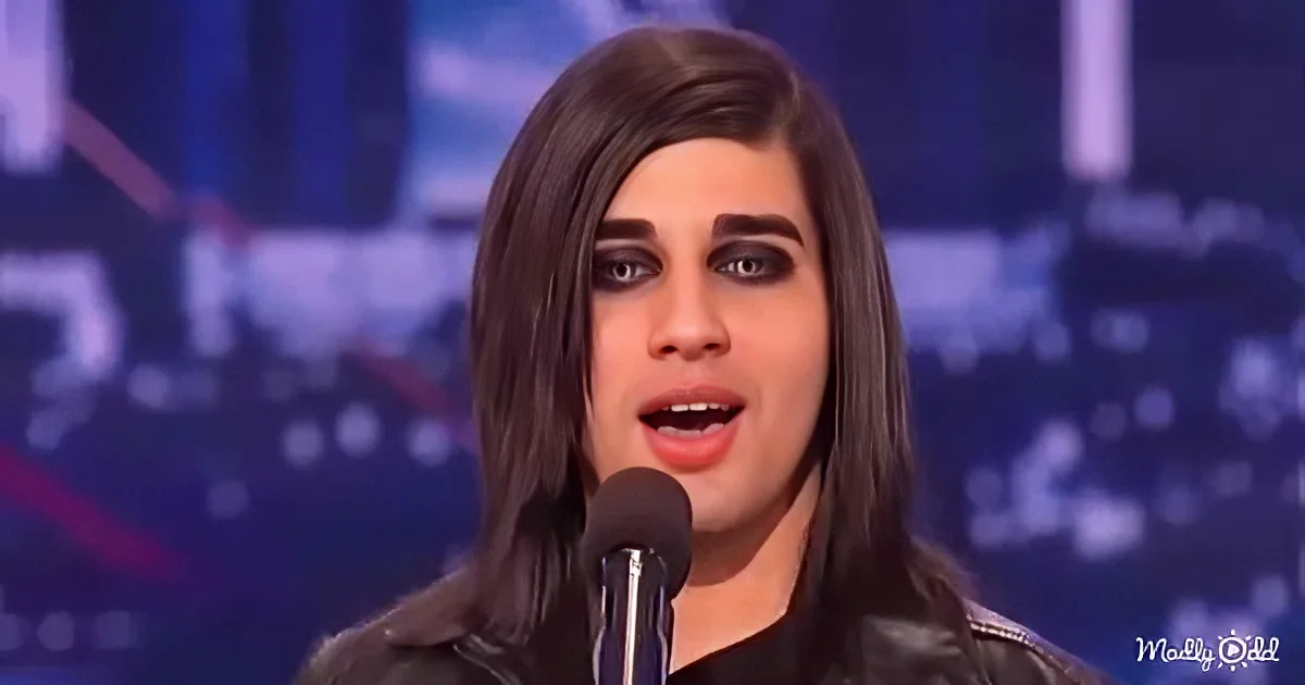 A person with long dark hair and makeup sings into a microphone on a stage with a blue background.