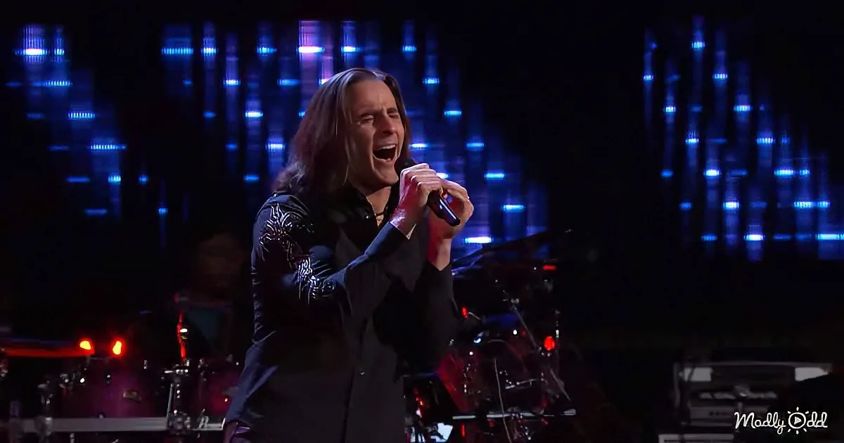 A man in a black outfit sings passionately on stage with a microphone, surrounded by blue lights and a band.