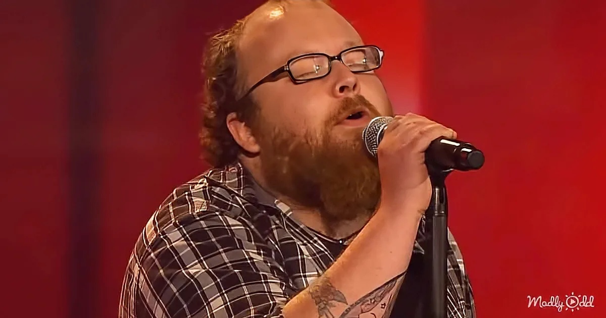 A man with glasses and a beard sings into a microphone, wearing a plaid shirt and a tattoo on his arm.
