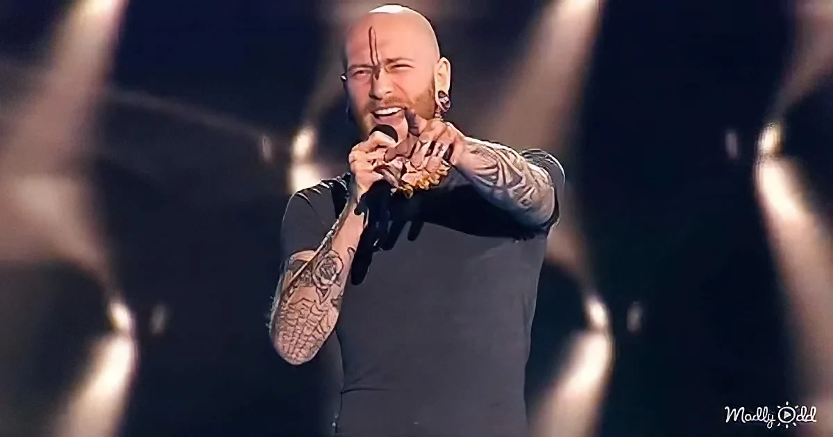 A man with tattoos sings passionately into a microphone on stage.