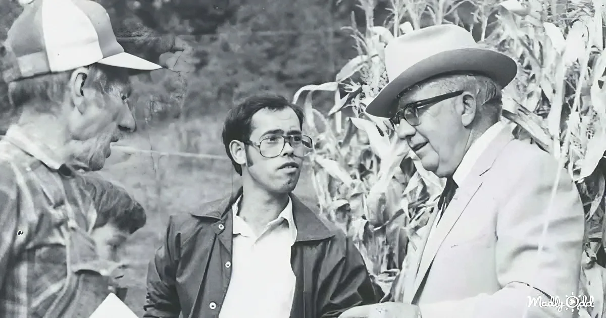 A man in a suit and hat is talking to another man in a field with corn stalks in the background.
