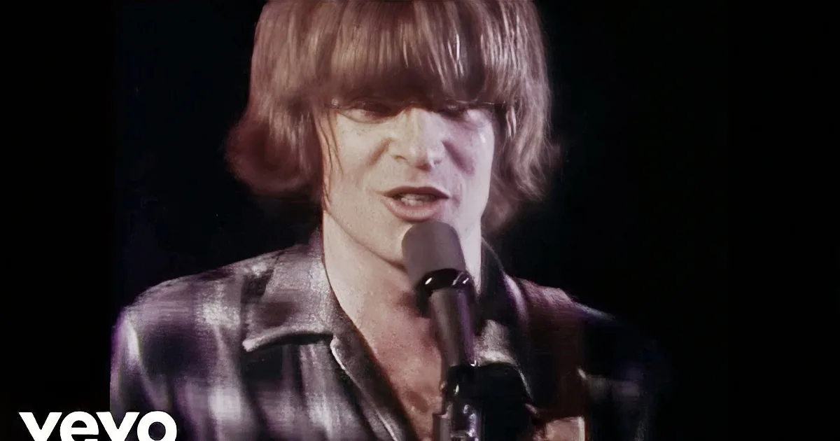 A person with shoulder-length hair is singing into a microphone, wearing a plaid shirt.
