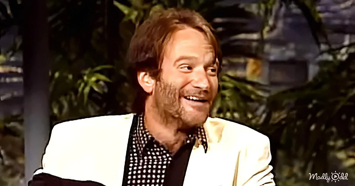 A man with a beard and mustache, wearing a white blazer and a black and white shirt, is smiling.