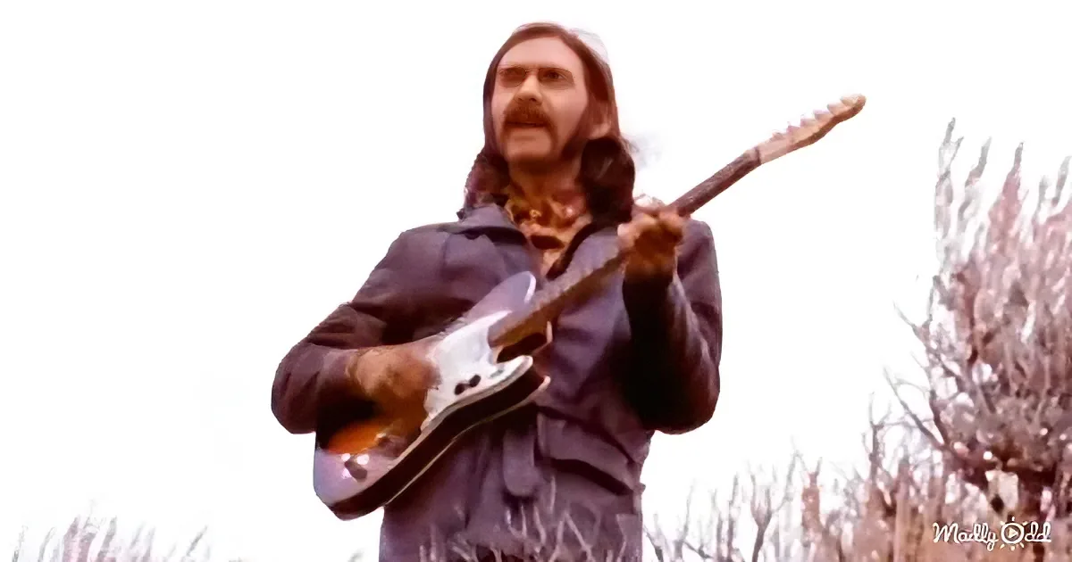 A man with long hair and a beard plays a guitar outdoors, surrounded by bare trees.