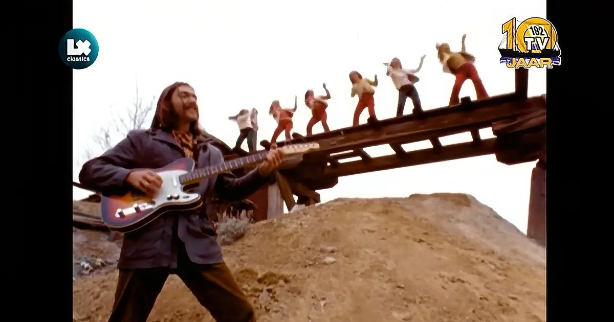 A man with long hair and a mustache plays a guitar, wearing a dark jacket and a patterned shirt.