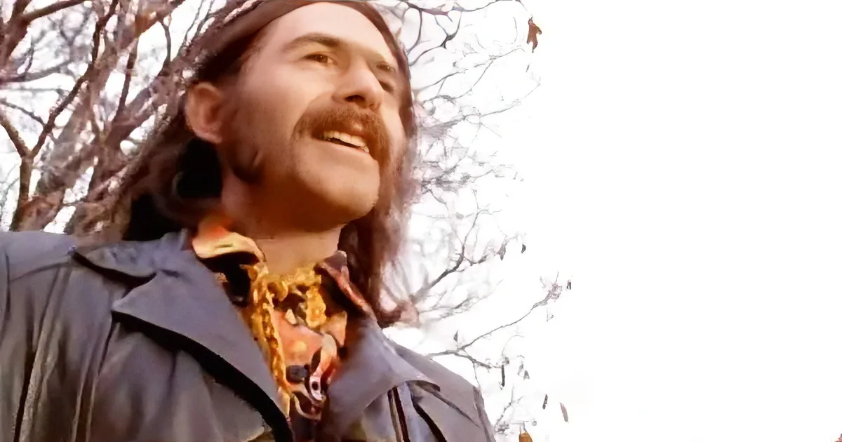 A man with long hair and a mustache smiles, wearing a dark jacket and a colorful scarf, with bare trees in the background.