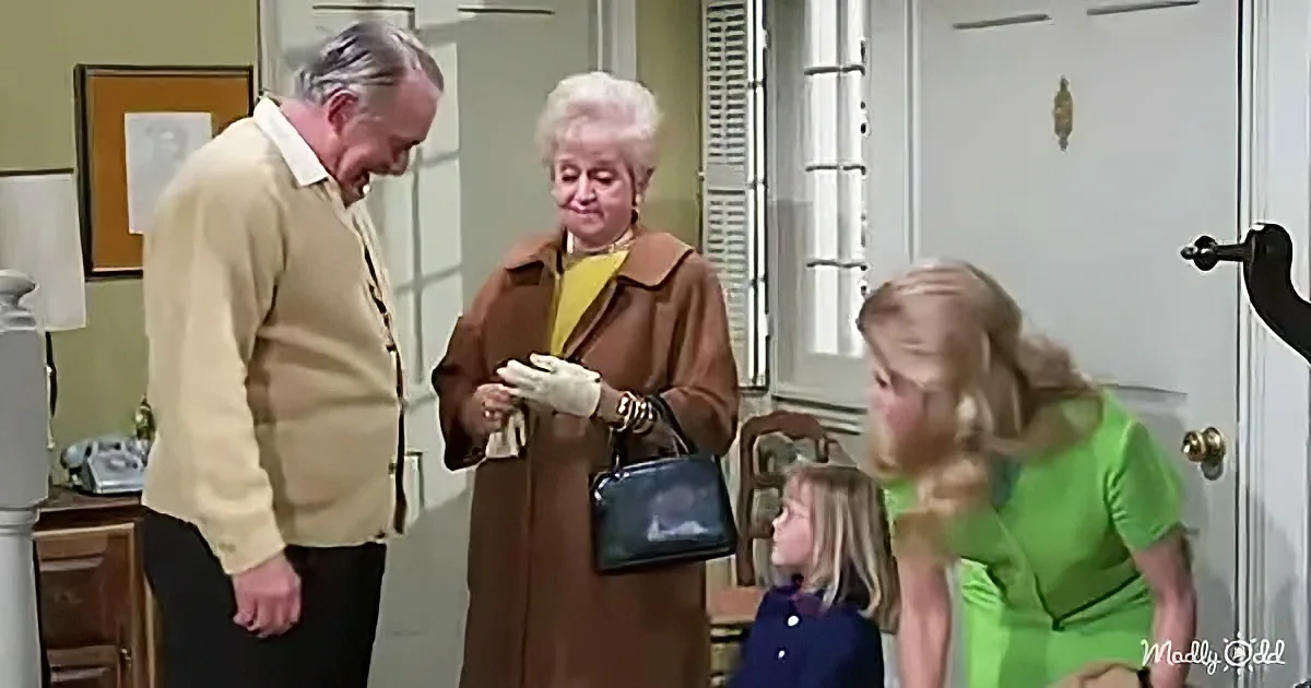 An older man in a yellow sweater interacts with a woman in a brown coat and white gloves, while a young girl and another...