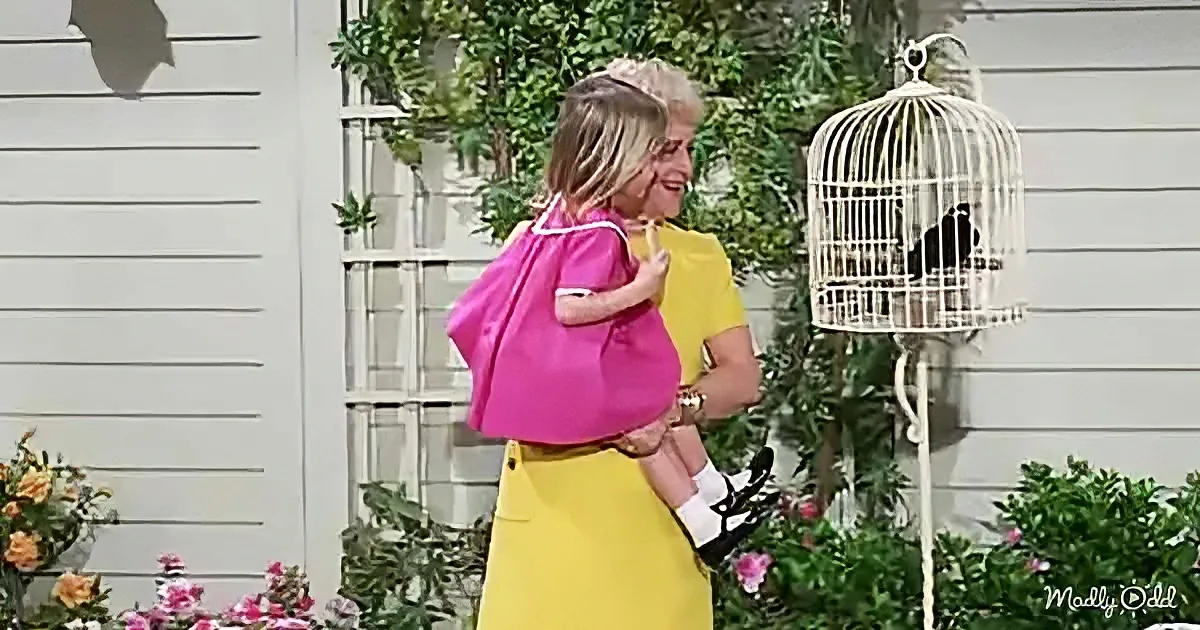 A woman in yellow holds a child in pink, standing near a white birdcage with a bird inside, surrounded by flowers and...