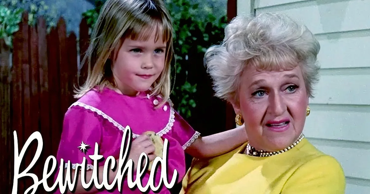 A young girl in a pink dress stands with an older woman in a yellow dress, both smiling outdoors.