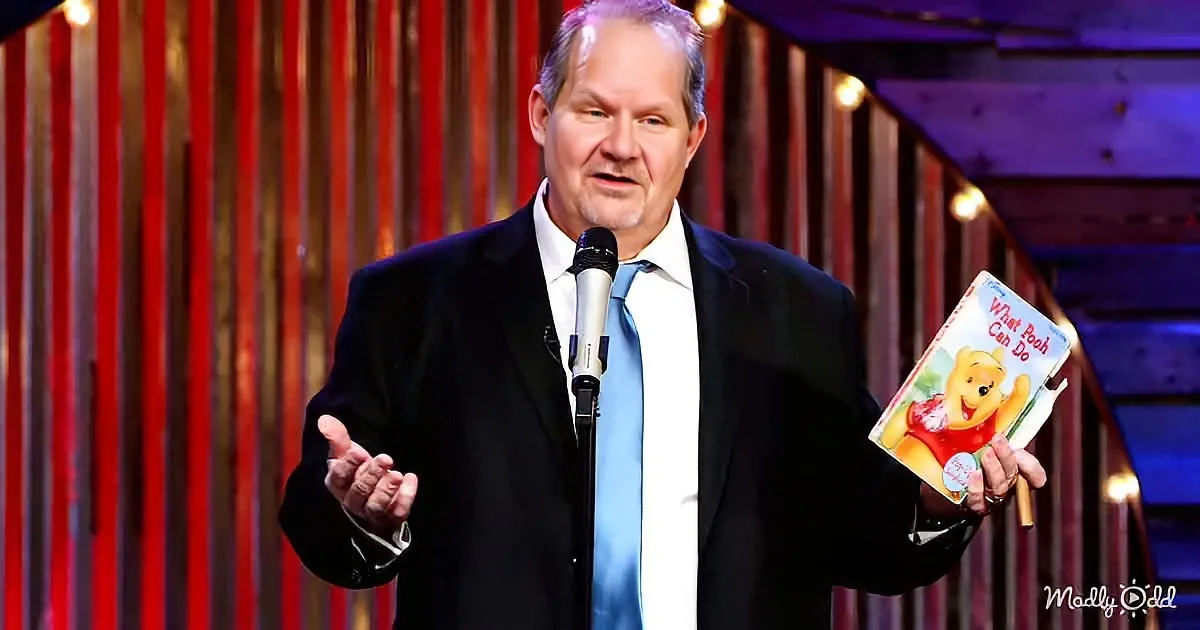 A man in a suit holds a "Winnie the Pooh" book while speaking into a microphone on stage.
