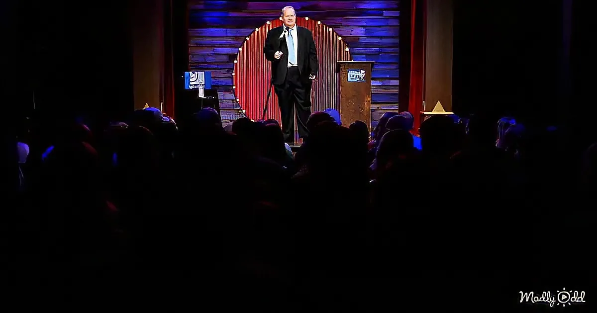 A man in a suit stands on stage with a microphone, addressing an audience in a dimly lit venue.