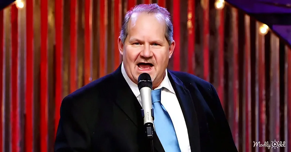 A man in a suit and tie stands on stage, holding a microphone, with a red curtain backdrop.