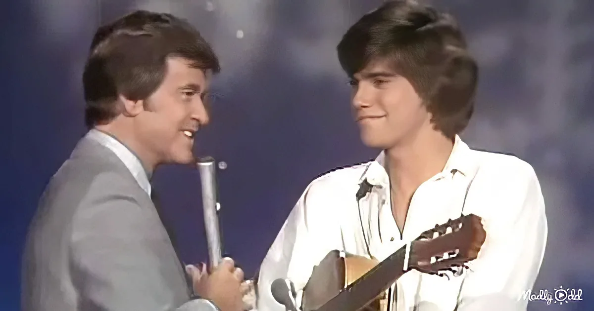 A man in a suit holds a microphone, smiling at another man playing a guitar.
