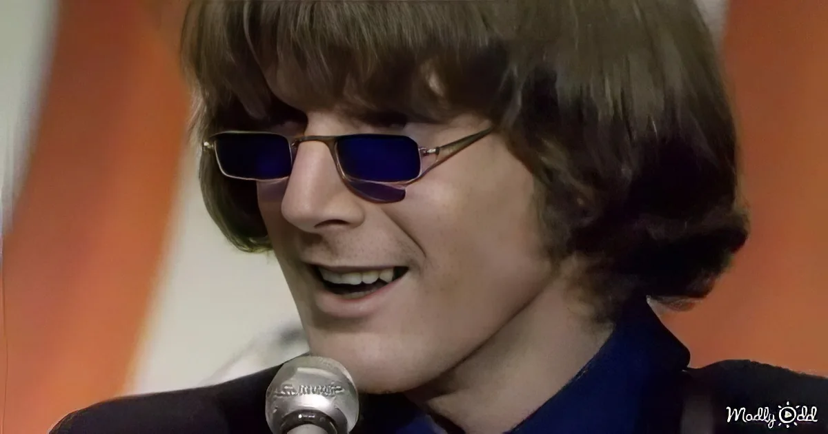 A person with shoulder-length hair and sunglasses smiles near a microphone against an orange background.
