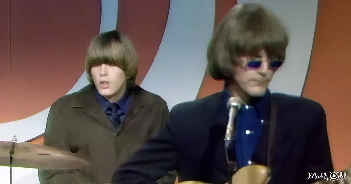 Two individuals in suits, one with sunglasses, seated at a table with microphones, against a colorful backdrop.