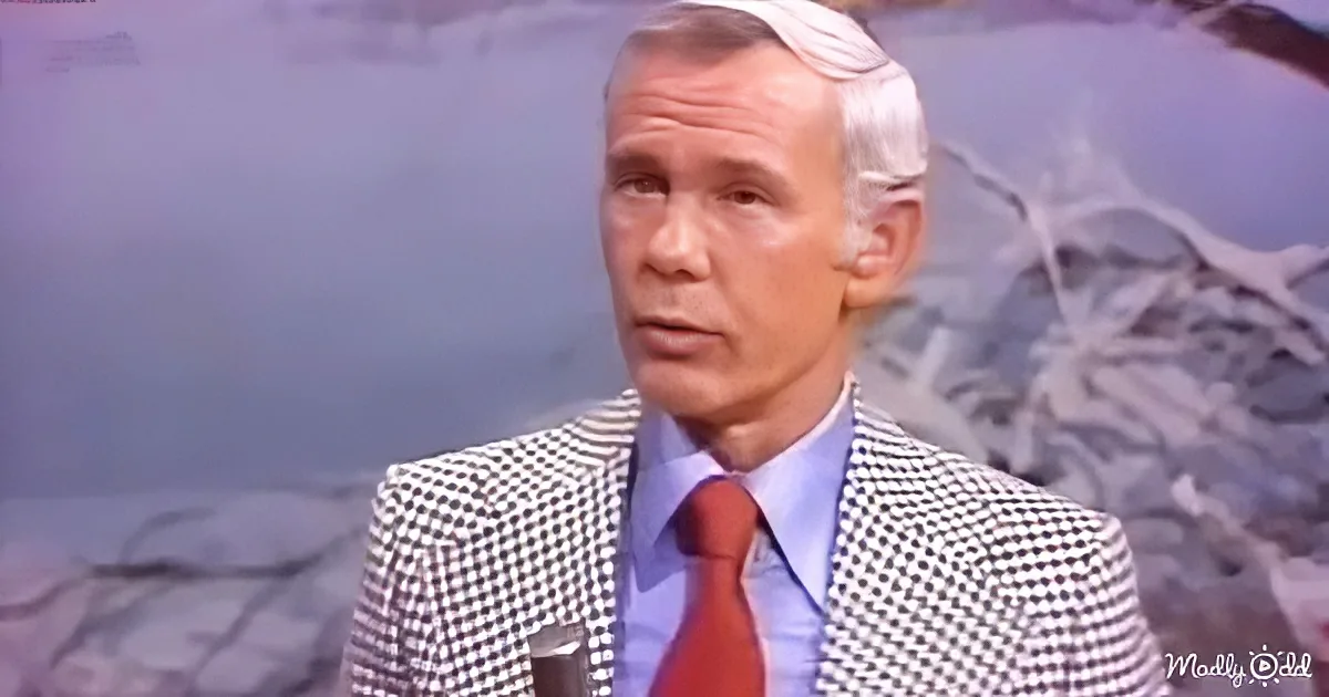 A man in a checkered suit and red tie is speaking, with a mountainous background.